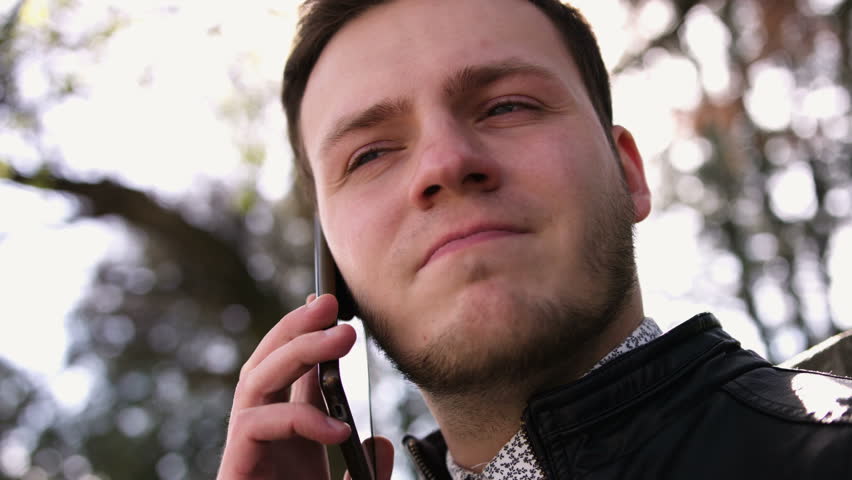 Portrait of an attractive young serious man with blue eyes looking toward speaks by phone in a sunny spring day in the park