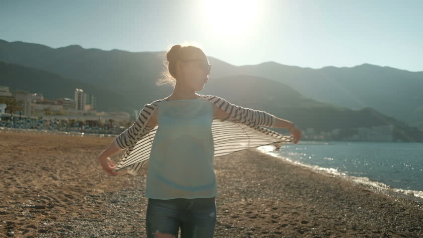 woman walking on sea shore, looking into distance, wind t-shirt. Dreaming female in ripped jeans, black striped sweater waving, sunglasses walks on beach, enjoying nature