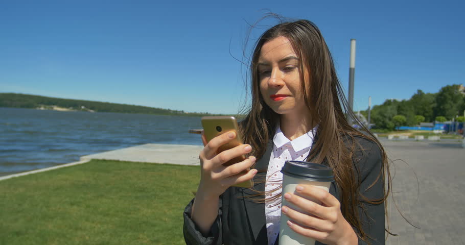 Woman browsing a cell phone and walking at the park. Drinking coffee at walking.