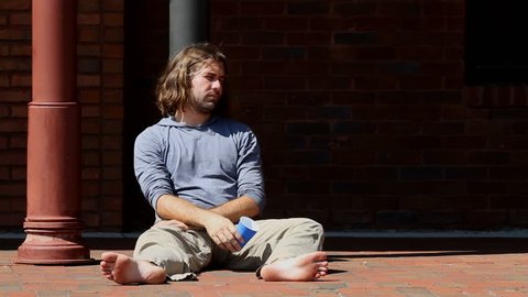 Intoxicated young adult man sits barefoot on the sidewalk leaning against a column holding his drink in a plastic cup.
