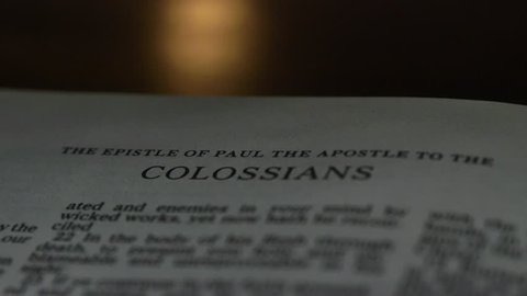The Bible-  A slow, shallow depth of field, candle lit pan across a page of the Bible that  reveals the heading for the book of Colossians.