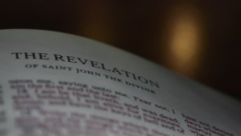 The Bible-  A slow, shallow depth of field, candle lit pan across a page of the Bible that  reveals the heading for the book of Revelation.