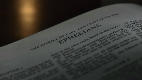 The Bible-  A slow, shallow depth of field, candle lit pan across a page of the Bible that  reveals the heading for the book of Ephesians.