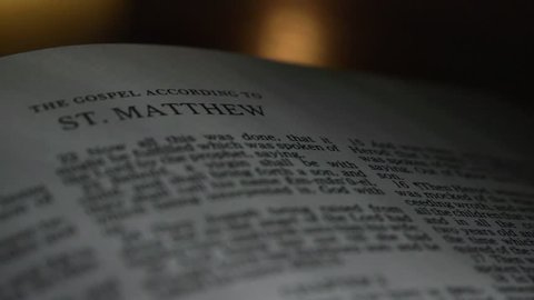 The Bible-  A slow, shallow depth of field, candle lit pan across a page of the Bible that  reveals the heading for the book of Matthew.