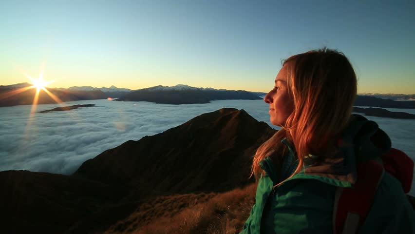 Young woman hiking stands on a mountain peak above the clouds and outstretches arms, sun shining over the mountains. Beautiful sunbeam effect making an idyllic landscape. Shot in New Zealand Royalty-Free Stock Footage #28800781
