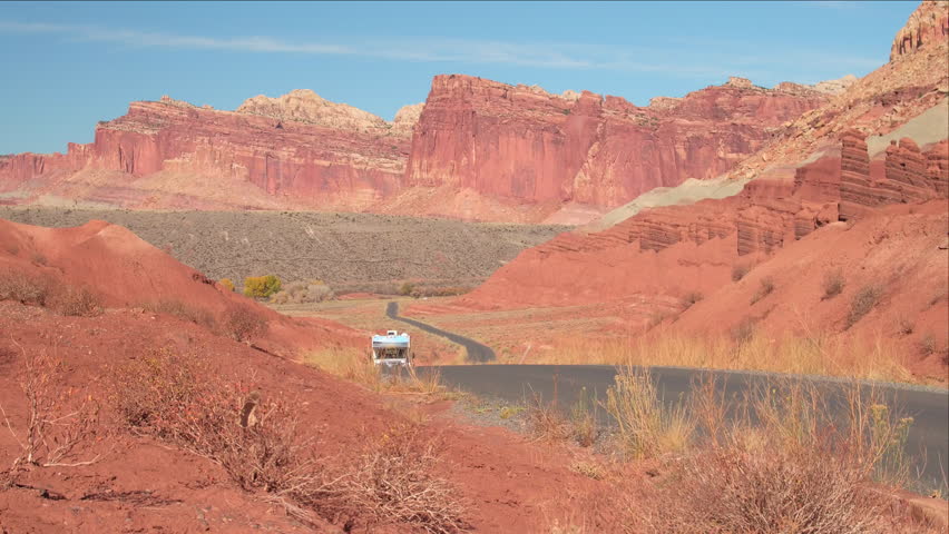 RV camper driving along empty road leading past butte and mesa mountains in red rocky desert on summer day. Trailer home traveling in Capitol Reef Canyon past rocky mountain formations in red desert Royalty-Free Stock Footage #28812064
