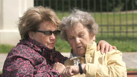 Two women sit and talk outdoors. One woman aged 80s dressed in yellow jacket looks at the results of physical activity using a wristband fitness tracker outdoors
