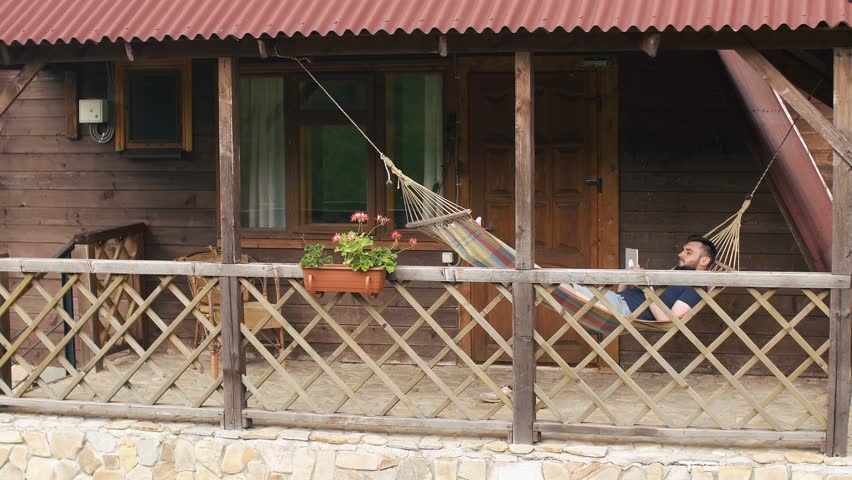 A young bearded man uses a tablet, lying in a hammock in a country house