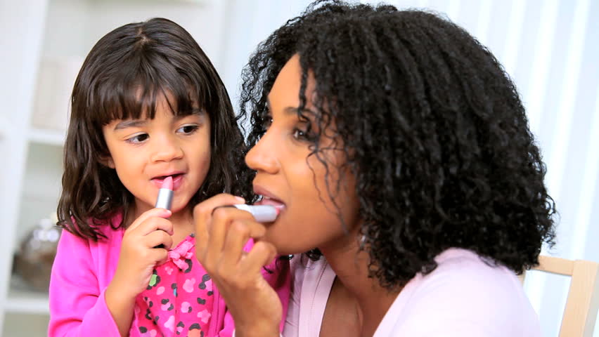 Loving African American mother with cute young daughter watching mom putting on lipstick