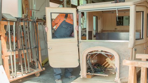 A middle-aged joiner is adjusting the door on the vintage wooden car. He is building a brand-new car in his workshop.