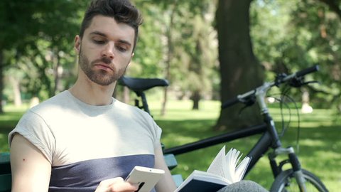 Handsome man answers cellphone while reading book in the park, steadycam shot
