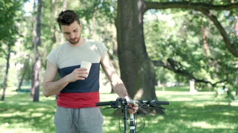 Relaxed man walking with his bicycle in the park and drinking coffee, steadycam shot
