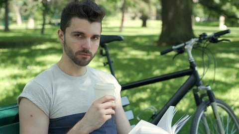 Handsome man smiling to the camera while drinking coffee and reading book, steadycam shot
