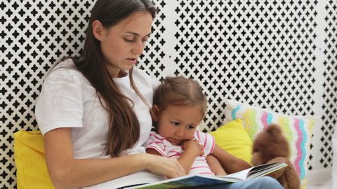 Young mother reading book for her little daughter. Mom and daughter sitting on the floor and learning. Domestic education concept.