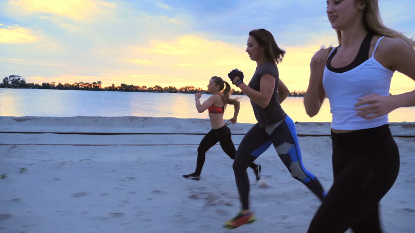 Three athletic women in a tracksuits are jogging along the sand pier of the cargo port, in the twilight of the morning dawn. sky with clouds, cargo cranes,
