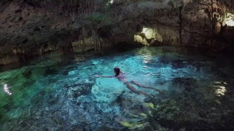 CLOSE UP Young woman swimming in turquoise pool in breathtaking underground cave. Tourists exploring a fascinating architecture of the stalactites and stalagmites in Aktun Chen cenote sinkhole, Mexico