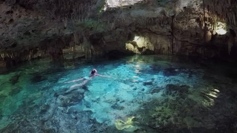 Young woman swimming in turquoise pool in magical underground cave with sunlight coming from the hole in the ceiling. Fascinating architecture of the stalactites and stalagmites in Aktun Chen cenote