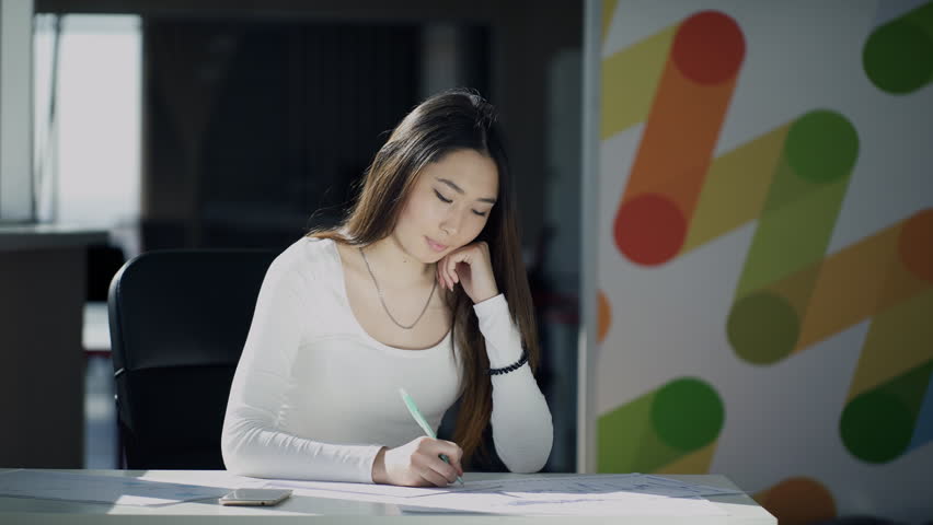 Young woman writes sitting at table indoors. Secretary makes notes on white sheets of documents lying in front of her on wooden desk in office of law company. Attractive brunette with long hair