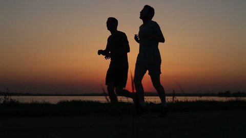 Two men jogging on lake shore sunset backlit silhouette. Twilight horizon sun.