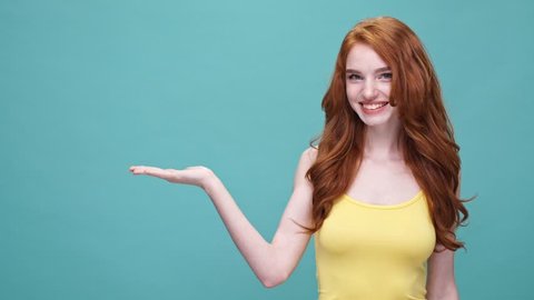 Happy cheerful redheaded girl presenting copyspace on her palm and winking isolated over blue background