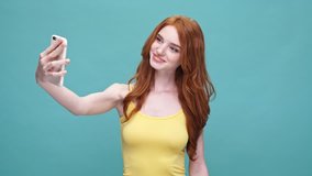 Happy pretty redheaded girl taking a selfie with mobile phone and making funny face while standing isolated over blue background