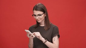 Smiling happy young girl in eyeglasses holding mobile phone and texting while standing isolated over red background