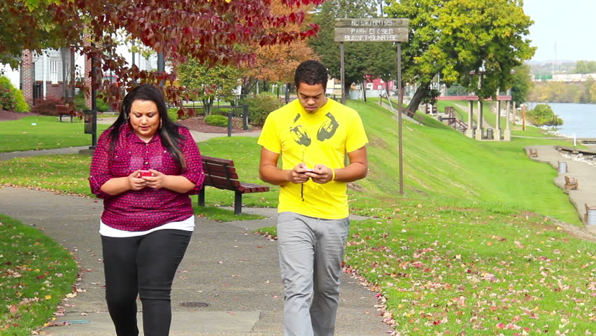 A young couple walks in the park while texting.