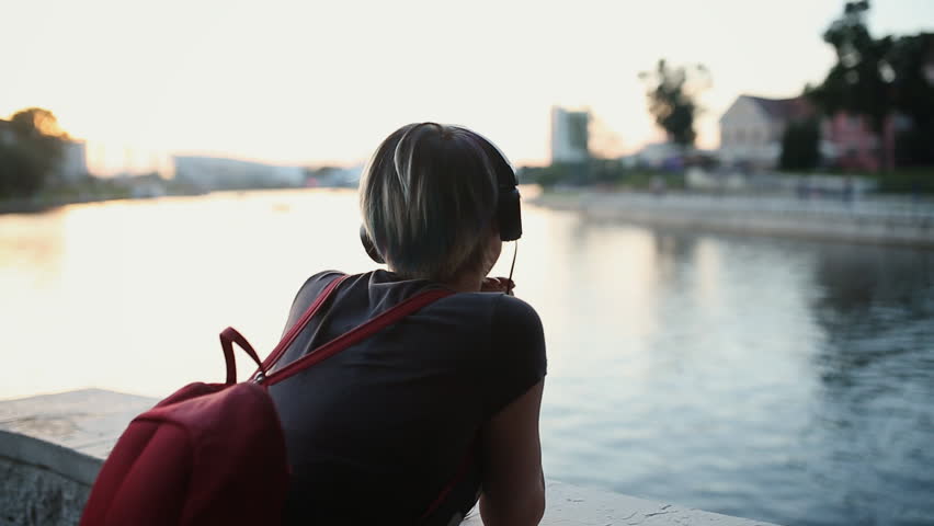 Young beautiful girl with a short haircut in headphones walking through the evening city and listening to music