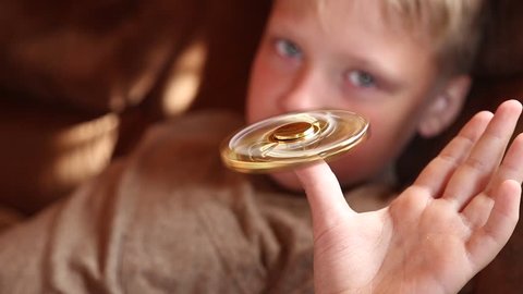 Little child playing with modern popular hand toy - spinner. Boy laying on brown cozy sofa in home interior and rotating golden anti stress spinner with one finger. Real time full hd video footage.