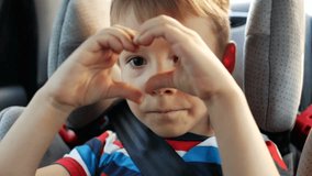 Little boys hands making heart shape gesture while sitting in car seat in summer evening. Young boy looking through the heart from hands to the camera. Child hands showing heart. Happy family concept