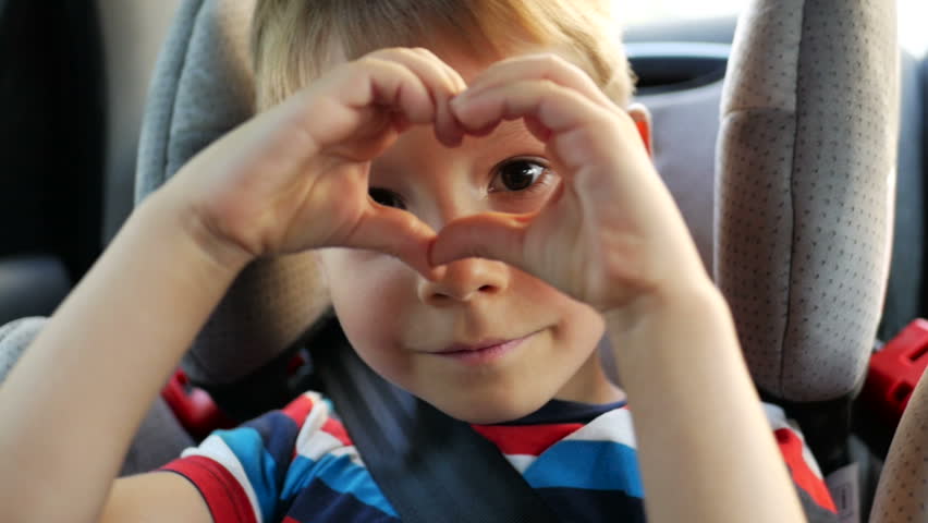 Little boys hands making heart shape gesture while sitting in car seat in summer evening. Young boy looking through the heart from hands to the camera. Child hands showing heart. Happy family concept