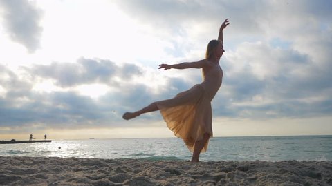 Beautiful scene of a dancing ballerina in beige silk dress and pointe on sand beach near ocean or sea at sunrise or sunset. Young blonde sensual woman with long hair practicing classic exercises.
