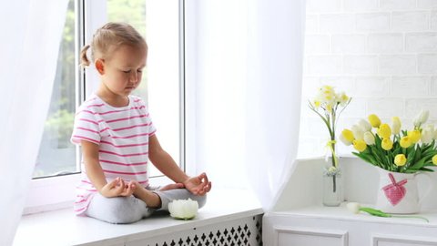 Tender video about little cute girl sitting in yoga pose and meditates on the window sill at light room with natural light. Child sitting in lotus pose with closed yeys.