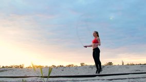Beautiful, slender young woman is engaged in fitness, jumping rope, At dawn, near a cargo crane, at the sandy quarry beach, in the sunlight, video shooting against the sun.