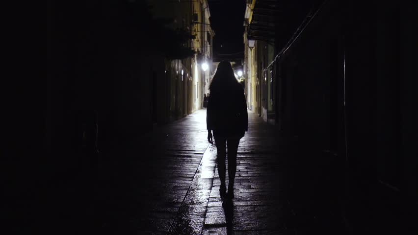 Woman silhouette walk on street of Pag city 4K. Static shot of person in focus in middle of frame walking towards lights in the end of street. Surrounded with black dark.