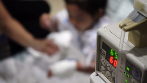 Close-up of infusion pump with parent and sick little child blurred in the hospital. Patient in the hospital is receiving a saline solution by dripping machine.