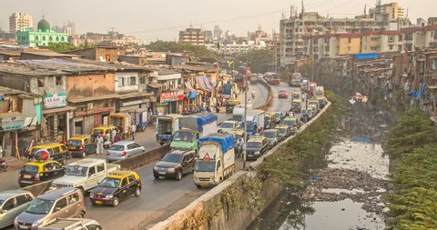 Time lapse shot of vehicles passing on a busy road through slums, while polluted nullah (canal) flowing alongside, Dharavi, Mumbai, India