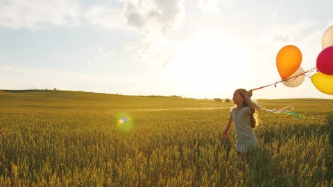 Little girl in cute dress running in the golden wheat field at sunset with balloons in the hand.