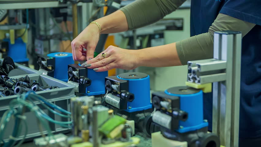 A young woman in the factory is putting the cover on the top of the item. She is working behind a conveyor belt.