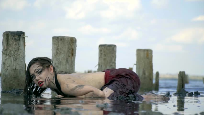 Beautiful blonde smeared in mud in a wet dress, lies in the middle of the estuary estuary, touching her face near the wooden posts from the ruined salt pool against the blue sky