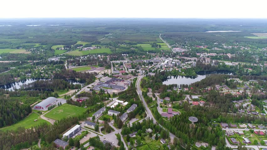 4K aerial view of small town with sport field, water tower, houses, lakes, and forest. Camera rising above looking towards horizon. Cloudy summer day at city of Suonenjoki, Finland. Scandinavia.