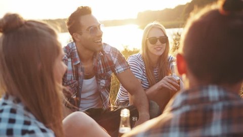 A company of four young friends having a picnic by the river, they are chatting, laughing, drinking beer. Casual wear, summertime, bright light of the sunset. Being happy, young and free.