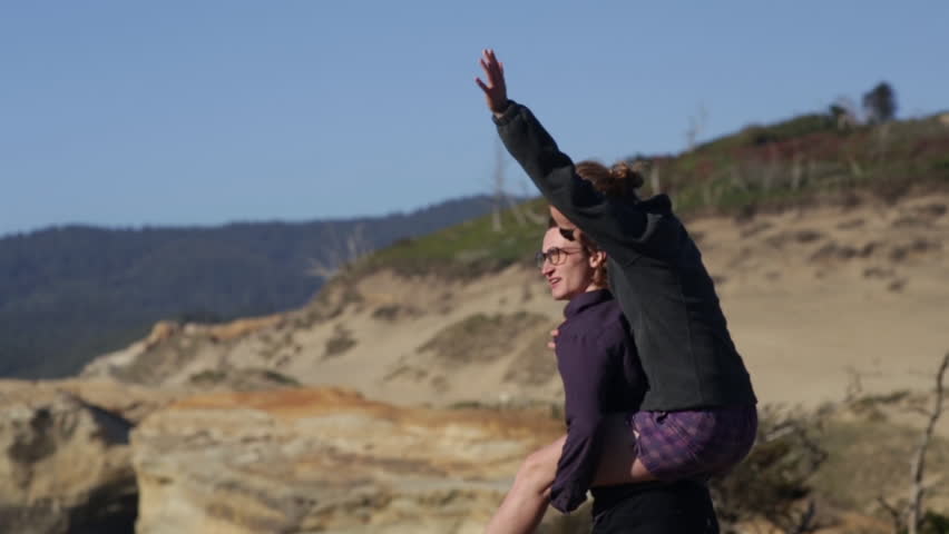 Man Gives Woman A Piggy Back Ride, She Puts Her Arms In The Air In Celebration, They Enjoy Ocean Views From Cliff