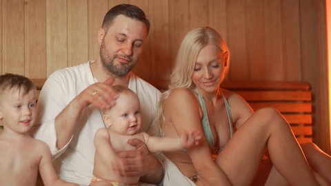 Family of four is basking in a wooden sauna. A young married couple with two children - a boy of 5 years and a one-year-old baby - sit in a sauna and have a good time together