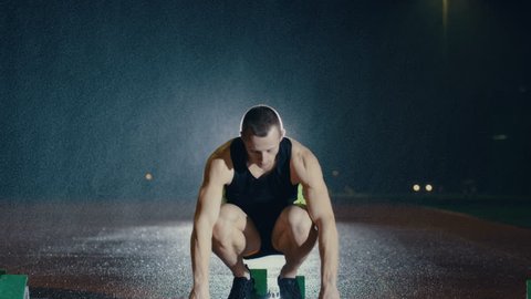 Male athlete training at running track in the dark & in the rain. Slow motion.
