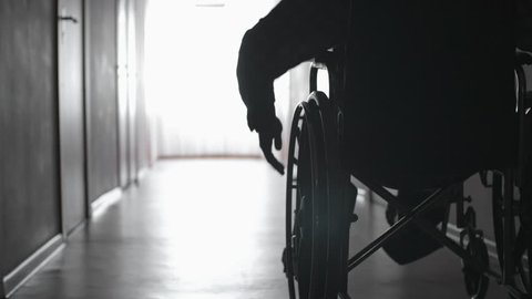 Tracking mid-section shot of silhouette of man turning wheels of wheelchair with hand and riding along dark corridor
