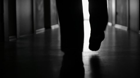 Tracking with low-section of silhouette of legs of businessman walking along dark hallway; black and white slow motion shot