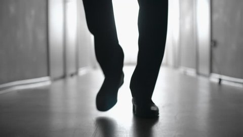 Dolly with low-section of silhouette of legs of man walking along hallway in slow motion; black and white shot
