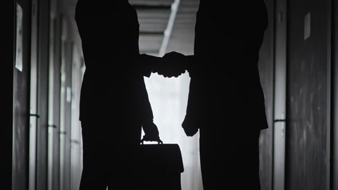 Silhouettes of unrecognizable businessman with briefcase and his business partner standing in dark hallway and exchanging handshake in agreement
