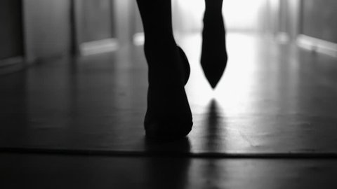 Dolly with low-section of silhouette of female legs in high heels shoes walking along corridor; black and white slow motion shot